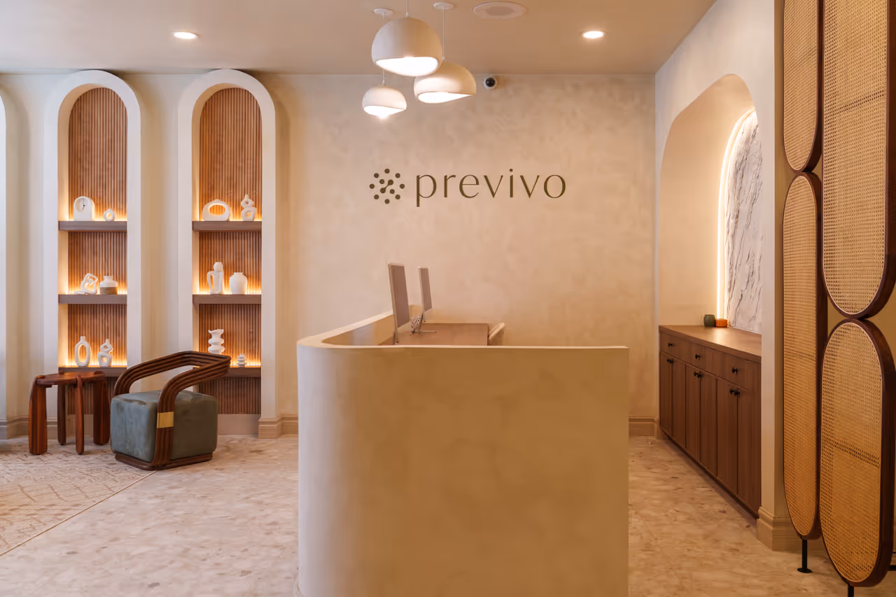 Minimalist reception area with curved beige desk, modern chairs, lit shelves, and Previvo logo on wall.