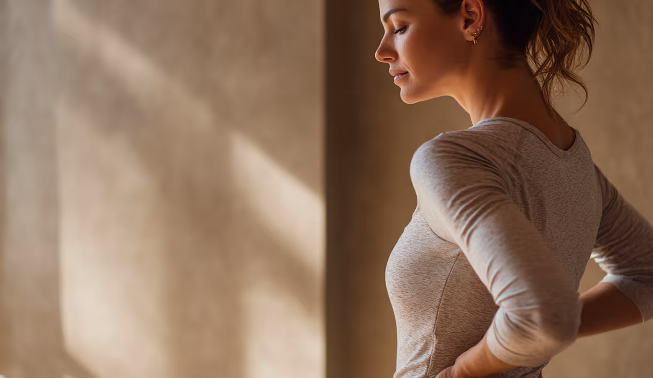 Woman in a gray shirt with hands on hips, eyes closed, standing against a beige wall.