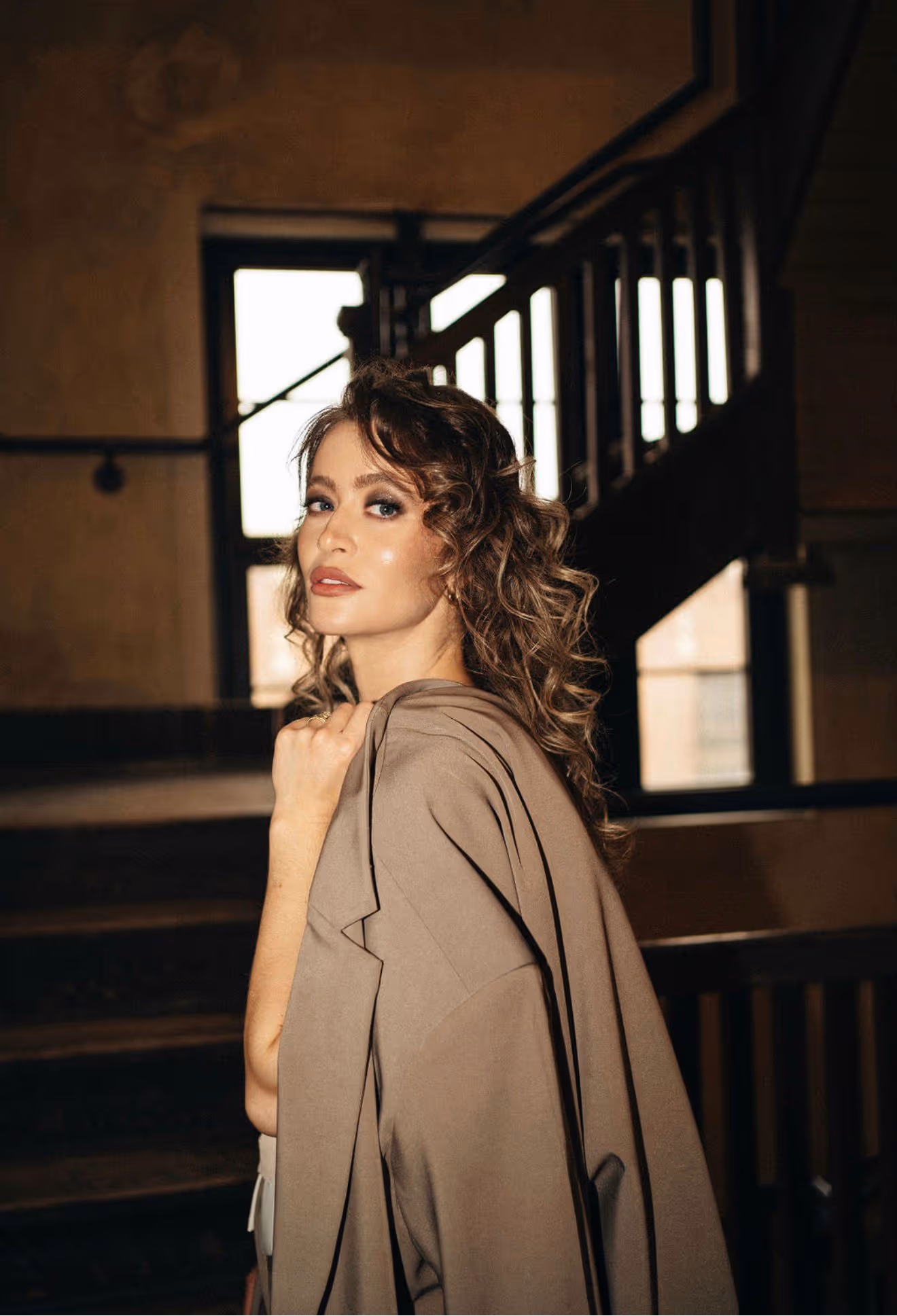 Woman with curly hair and makeup wearing a brown coat over her shoulder indoors by stairs.