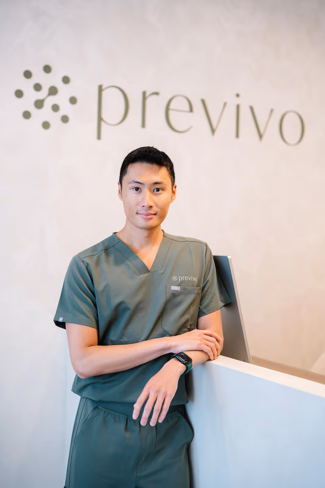 Young male healthcare worker in green scrubs standing with arms crossed by a reception desk with Previvo sign.