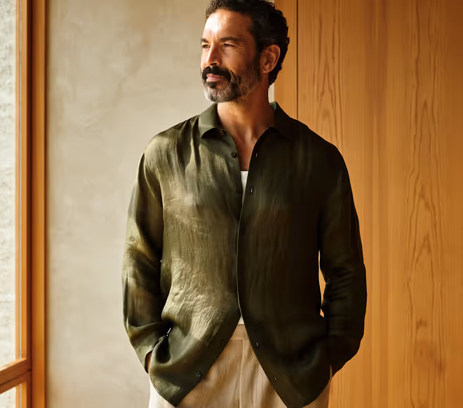 Man with beard wearing a green shirt and beige pants standing with hands in pockets looking out a window.