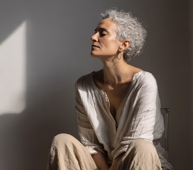 Woman with curly gray hair sitting pensively in soft light against a neutral wall.