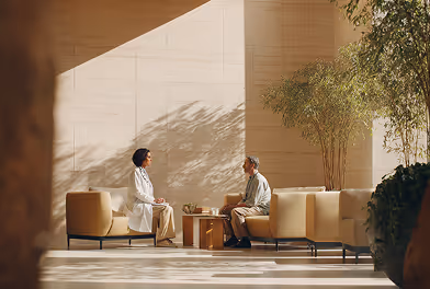 Two people sitting and talking in a sunlit modern room with beige furniture and plants.