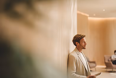Man in a beige blazer leaning against a wall holding a tablet in a softly lit modern room.