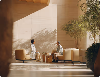 Two people seated facing each other in a sunlit modern room with plants and beige furniture.